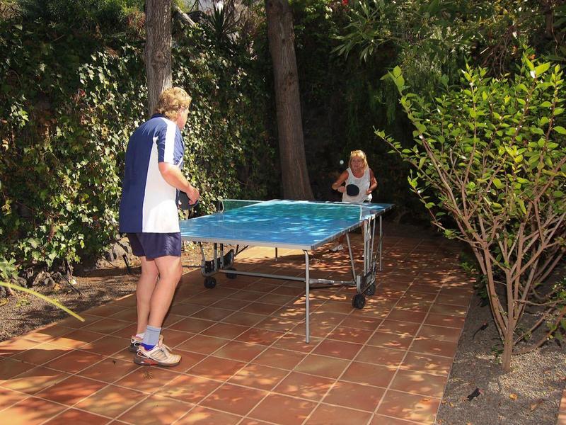 Two people play table tennis on an outdoor terrace surrounded by trees.