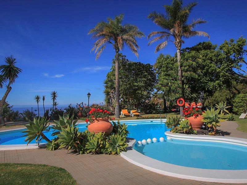 Recreational pool area with blue water slide, surrounded by palm trees and colorful flowers under clear sky.