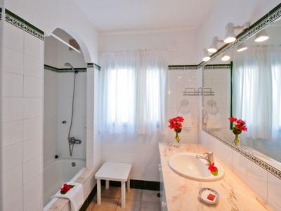 Helles Badezimmer mit Marmorwaschbecken, großem Spiegel, weißen Fliesen und roter Blumendeko.