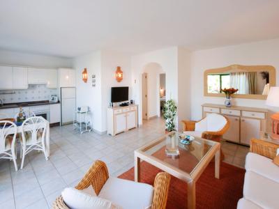 Helles Wohnzimmer mit weißen Möbeln, Terrakotta-Teppich, Glastisch und offener Küche im Hintergrund.