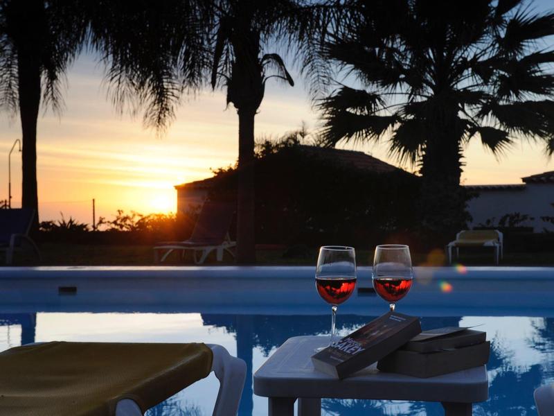 Two wine glasses on a table by a pool at sunset with palm trees in the background.