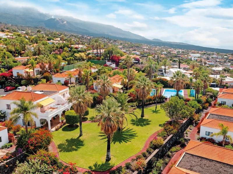 View of a hotel complex with green gardens, palm trees, and adjacent buildings under a blue sky.