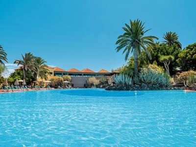 Großer blauer Pool mit Palmen, Sträuchern und Gebäude im Hintergrund unter klarem Himmel.
