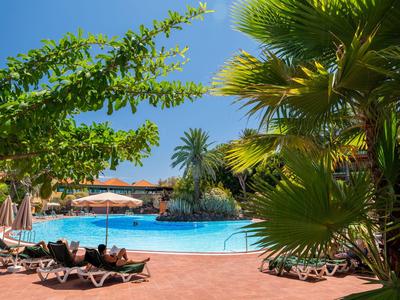 Pool area with lounge chairs, umbrellas, and tropical plants under a blue sky.