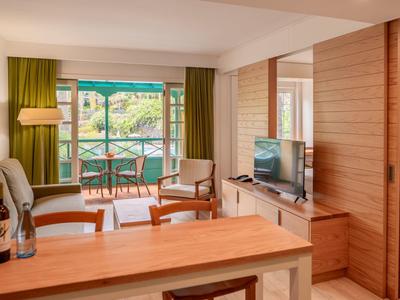 Modern eingerichtetes Wohnzimmer mit Holzdetails, Fernseher, Pflanzen und Blick auf Balkon mit Stühlen.