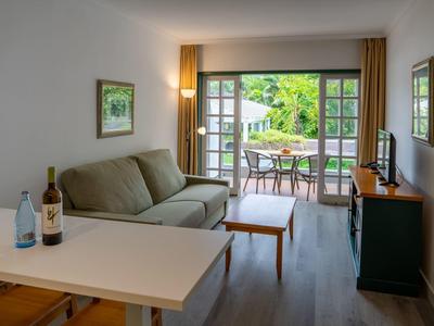 Helles Wohnzimmer mit grauem Sofa, Holztisch, TV, Balkon mit Stühlen und grünem Außenblick.