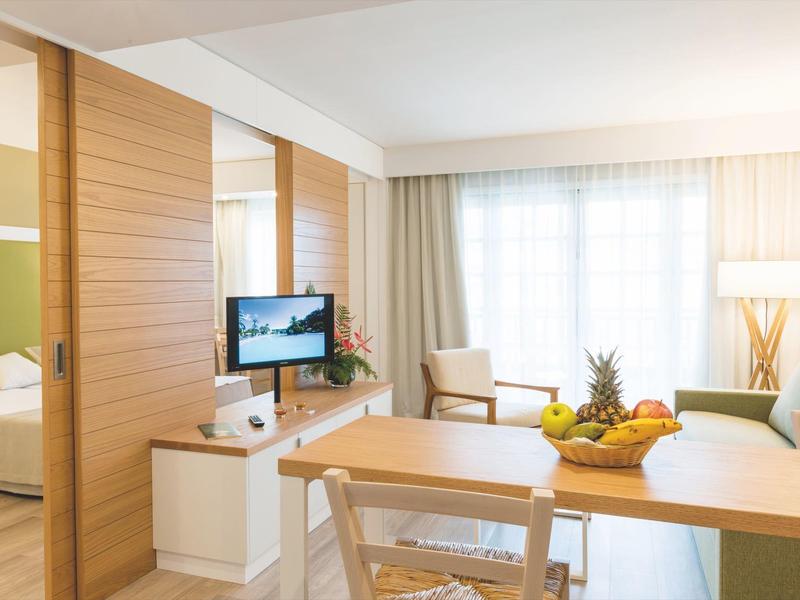 Habitación de hotel luminosa con mesa, sillas, sillones y TV junto a un dormitorio adyacente.