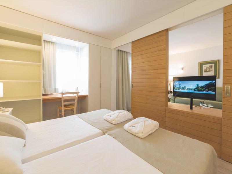 Habitación de hotel luminosa con dos camas, muebles de madera y televisor en la pared.