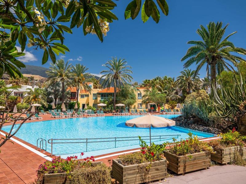 Gran piscina con sombrillas, rodeada de palmeras y casas de vacaciones bajo cielo despejado.