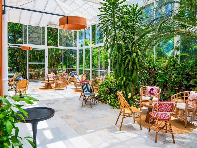 Luminosa sala de hotel con techo de cristal, plantas y cómodas áreas de asiento de mimbre.