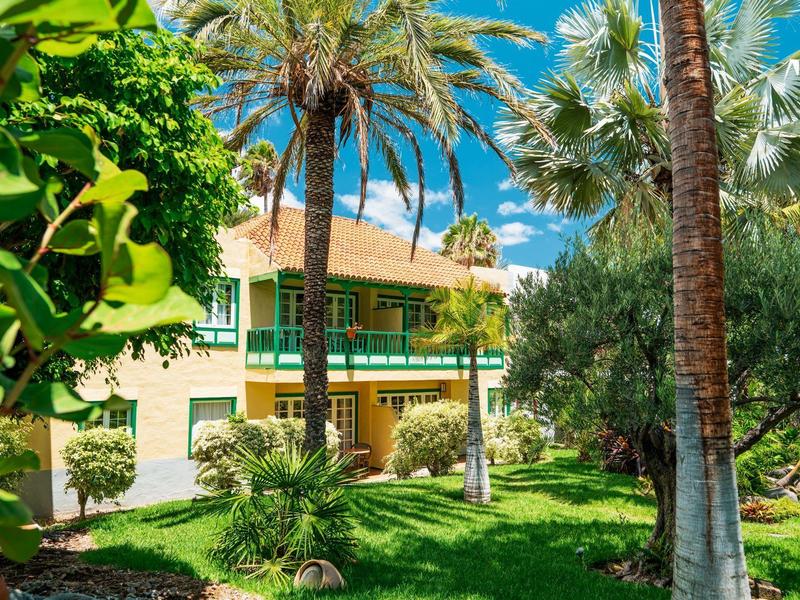 Edificio de hotel con terraza rodeada de palmeras y jardín exuberante bajo cielo azul.