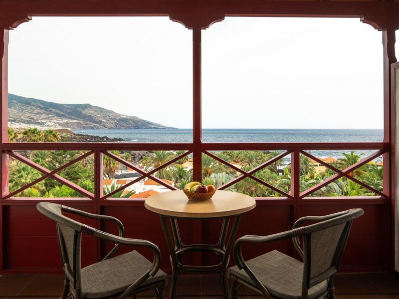 Balcón con mesa y dos sillas con vista al mar y montañas al fondo.