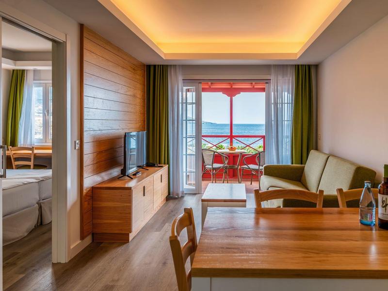 Sala de estar con muebles de madera, sofá, TV y balcón con vistas al mar y cielo al atardecer.