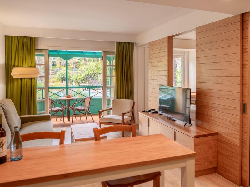 Habitación de hotel luminosa con balcón, cortinas verdes y mobiliario de madera, vista a un paisaje verde.