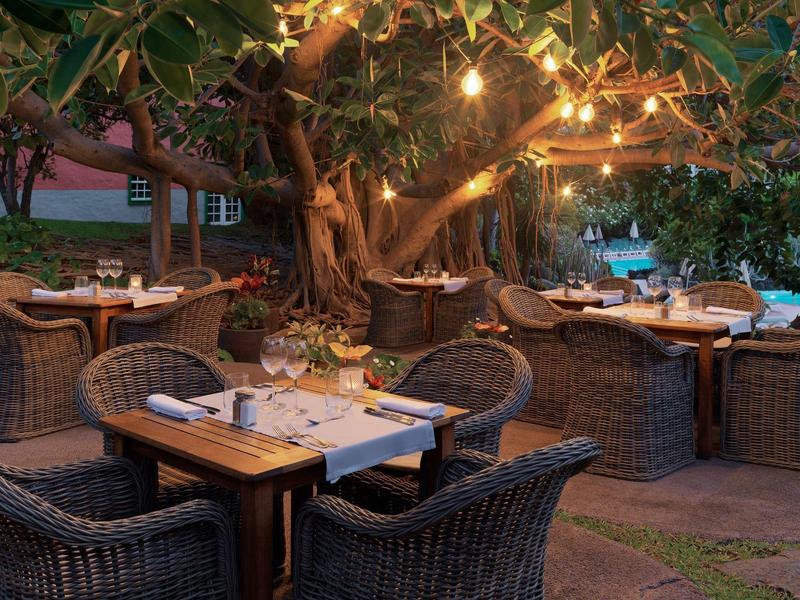 Acogedora zona al aire libre de restaurante con muebles de ratán y luces cálidas en el árbol.