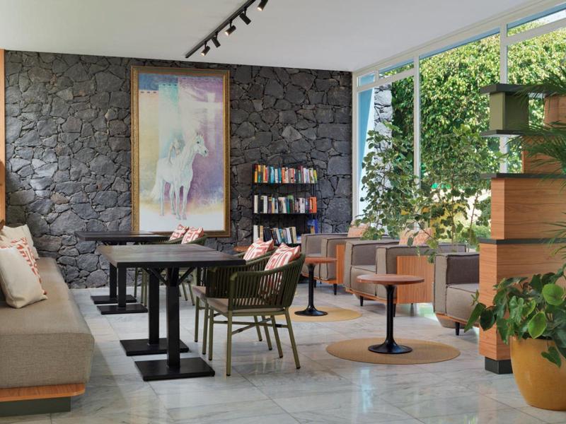 Área de salón de hotel decorada modernamente con asientos, estanterías y grandes ventanas.