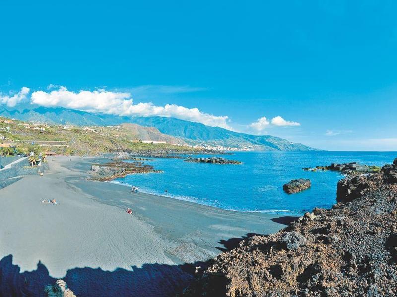 Playa con rocas volcánicas negras y mar azul tranquilo bajo cielo azul.