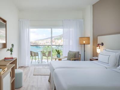 Lichte hotelkamer met groot bed, stoelen en balkon met uitzicht op zee.