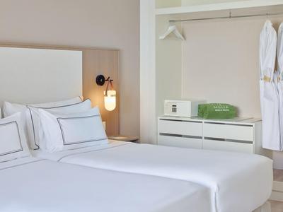 Lichte, moderne hotelkamer met twee witte bedden, nachtkastje, kasten en badjassen.