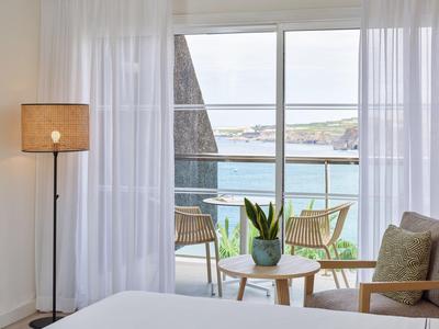 Lichte hotelkamer met grote ramen, uitzicht op terras en zee, vitrage en modern meubilair.