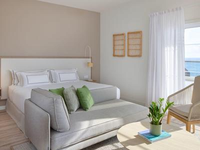 Heldere hotelkamer met tweepersoonsbed, bank, fauteuil en zeezicht door groot raam.