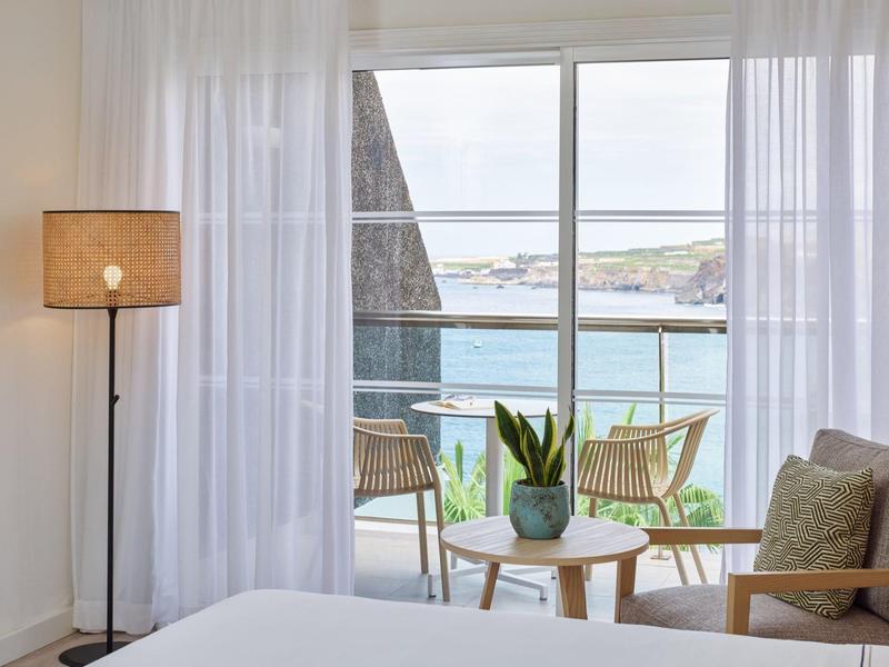 Lichte hotelkamer met grote ramen, uitzicht op terras en zee, vitrage en modern meubilair.