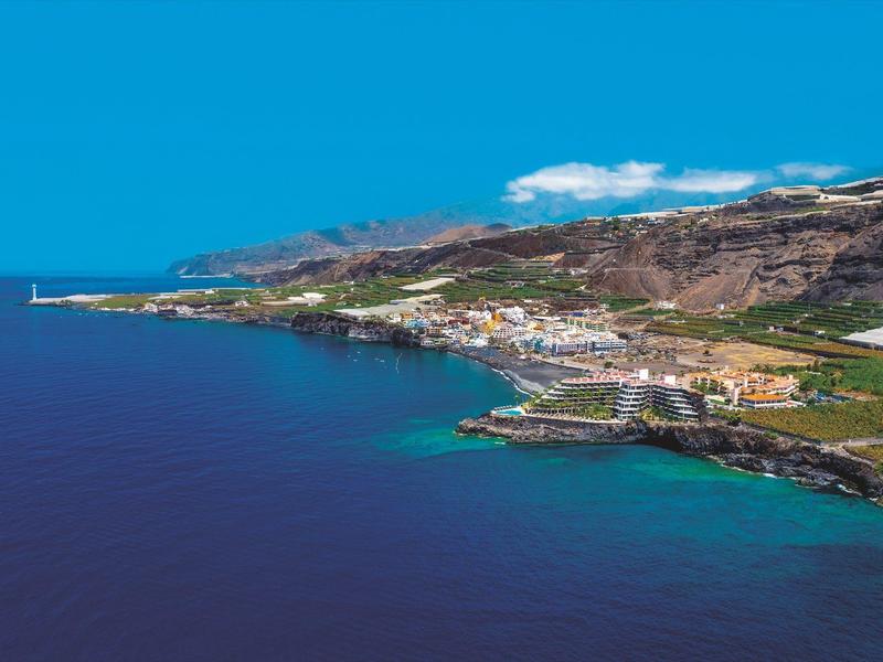 Kustplaats met helder blauw water, gebouwen en heuvels onder een blauwe lucht.