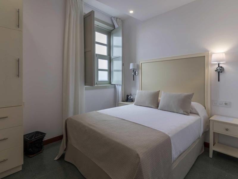 Schlichtes Schlafzimmer mit Bett, hellen Wänden, Fenster, Nachttisch und Schrank in neutralen Tönen.