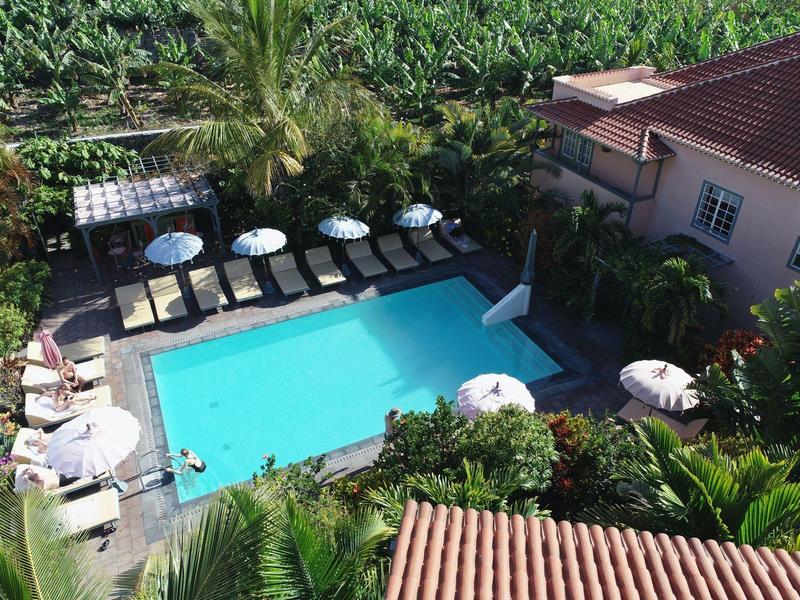 View of a rectangular hotel pool with sun loungers and umbrellas surrounded by tropical vegetation.