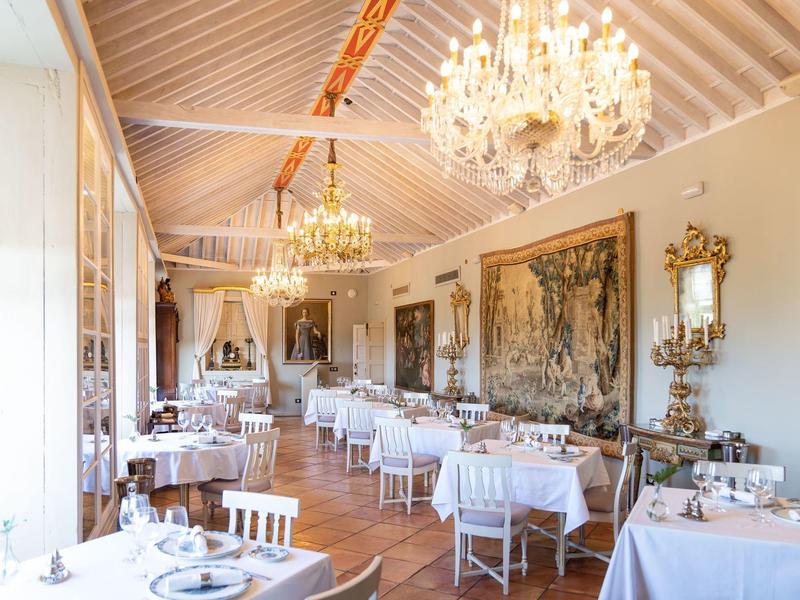 Elegant restaurant with white tablecloths, chandeliers, and tapestries in a bright room.