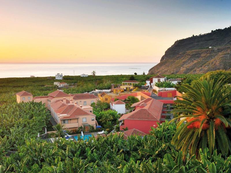 View of a hotel resort with red roofs surrounded by green vegetation and a hill in the background at sunset.