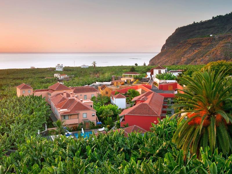 Small village with red roofs, surrounded by green fields and palm trees near a hill at sunset.