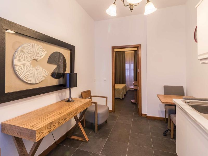 Moderner kleiner Raum mit Holzkonsole, Stuhl, Tisch, Küche rechts und Tür zum Schlafzimmer.