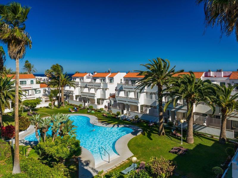 Complejo hotelero con amplia piscina, palmeras y edificios blancos bajo cielo azul despejado.