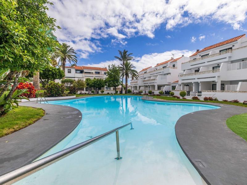 Gran piscina al aire libre con apartamentos blancos adyacentes bajo cielo azul y palmeras.