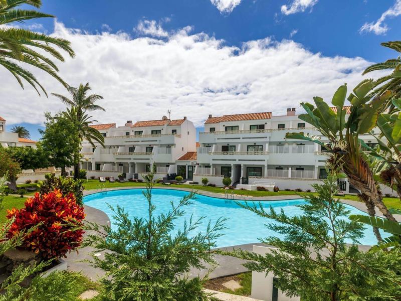 Complejo de apartamentos con piscina al aire libre, rodeado de vegetación tropical y edificios blancos.