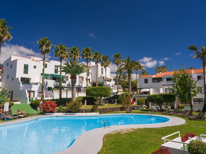 Piscina redonda frente a edificios blancos con palmeras y cielo azul claro.