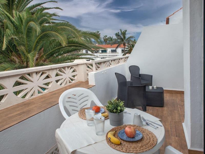 Balcón con mesa blanca, sillas, planta y desayuno con palmeras de fondo.