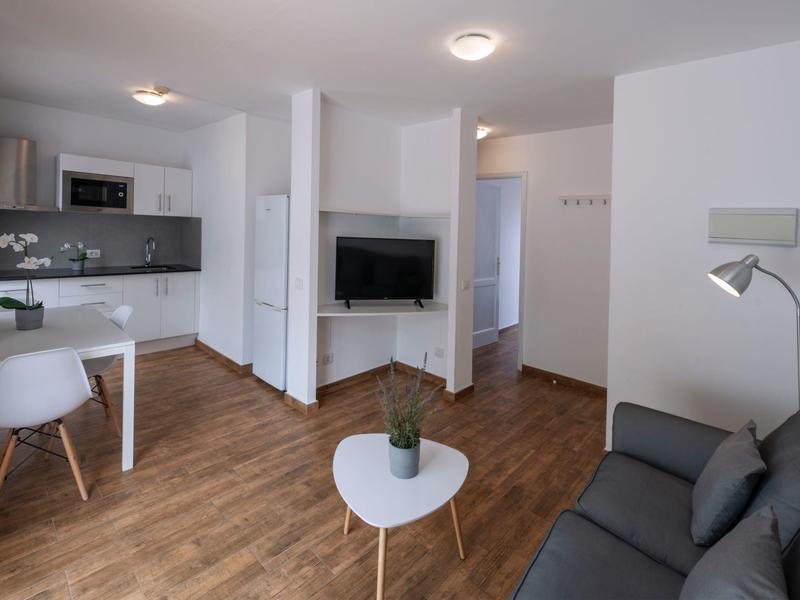 Área de estar moderna con sofá gris, mesa blanca pequeña y cocina abierta con suelo de madera