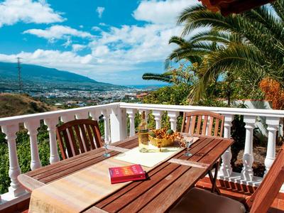 Terrassenbereich mit Holztisch und Stühlen, bunter Obstschale, Buch, Palmen und Bergsicht am Horizont.