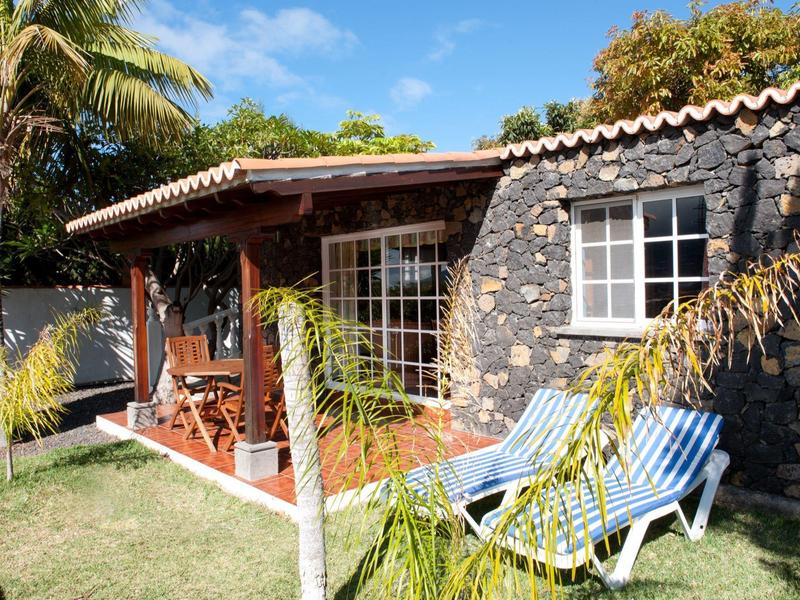 Casa de vacaciones con terraza, muebles de madera y tumbonas junto a césped y palmeras.