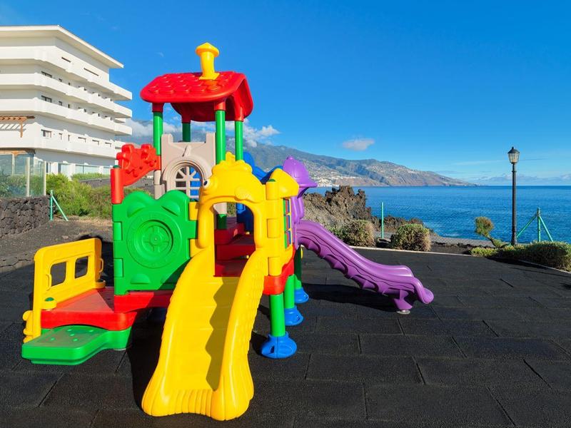 Colorida área de juegos infantiles frente a un hotel con vista al mar y cielo azul.