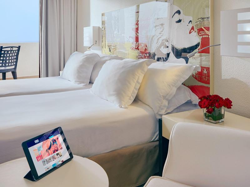 Habitación de hotel con dos camas, mesita, tablet y vista a una terraza con silla.