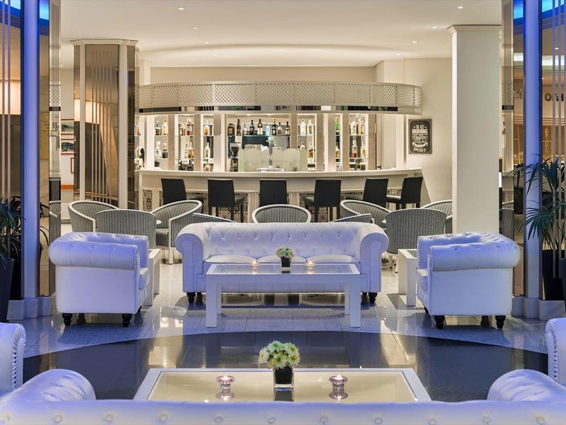 Área elegante de salón de hotel con sofás blancos y mesa de cristal central, iluminado modernamente.