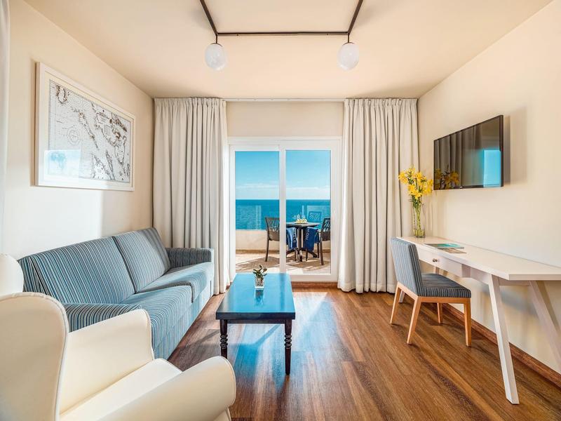 Sala luminosa con sofá azul, suelo de madera y vista al mar a través de ventana con cortinas.