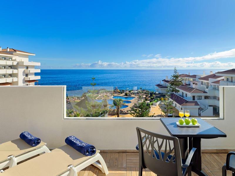 Terraza con mesa, dos sillas y tumbonas con vista al mar y edificios blancos bajo cielo azul claro.