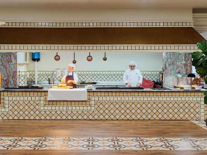 Cocina abierta de hotel con un chef detrás de un mostrador revestido de azulejos y suelo decorativo.