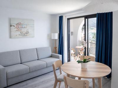 Helles Hotelzimmer mit grauem Sofa, rundem Holztisch, Stühlen und Balkon mit Meerblick.