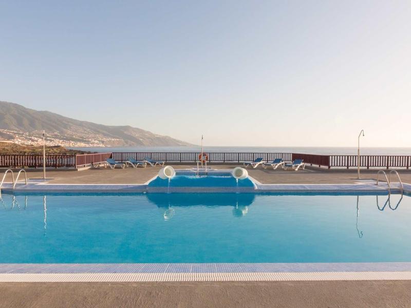 Empty outdoor pool with clear water overlooking the sea and mountains in the background.