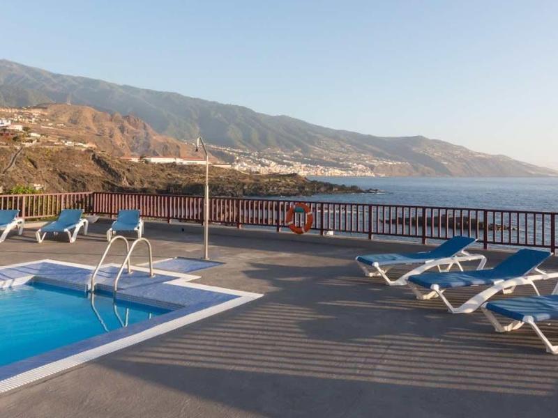 Pool area with lounge chairs and view of mountains and sea under clear sky.